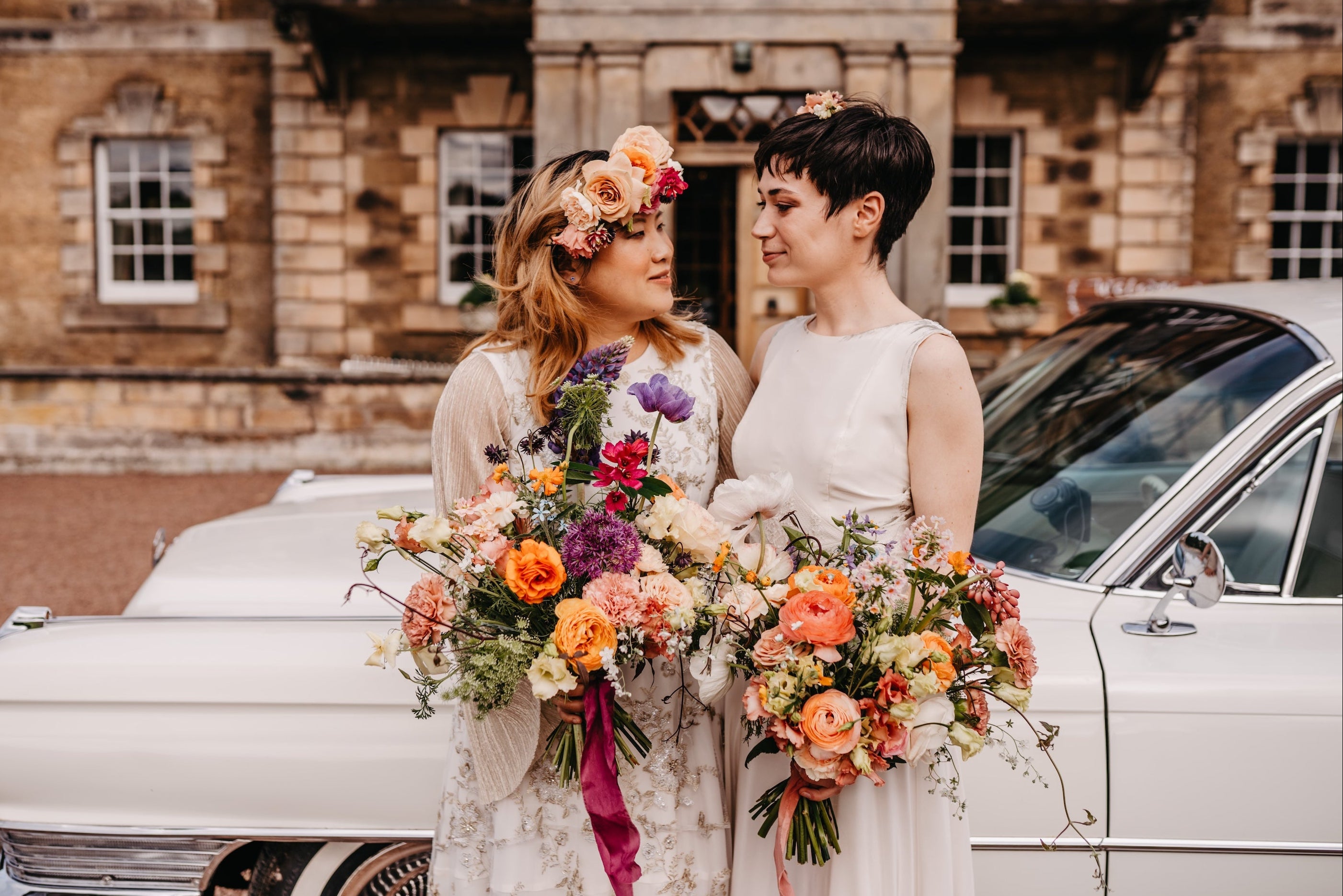 Two women standing in front of a vintage car with floral bouquets, wearing white dresses.