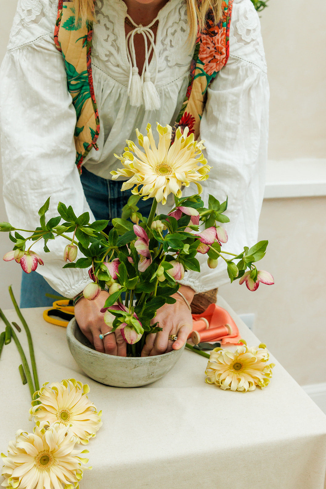 Person arranging flowers in a pot on a table with a colorful scarf draped over shoulders.
